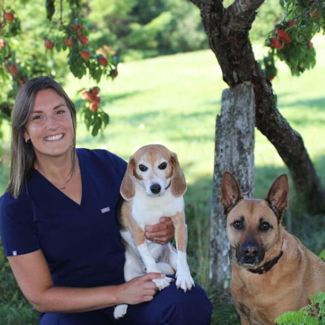 A woman holding a dog and sitting next to a tree.